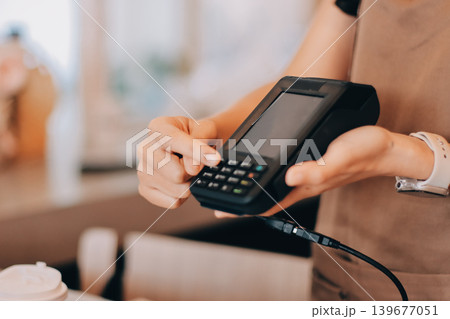 Friendly Asian female barista receives a credit card from a customer for payment. Both women are smiling in a warmly lit coffee shop with a rustic interior. Cashless payments. Small family business 139677051