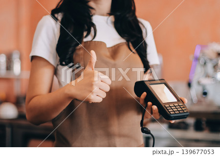Friendly Asian female barista receives a credit card from a customer for payment. Both women are smiling in a warmly lit coffee shop with a rustic interior. Cashless payments. Small family business 139677053