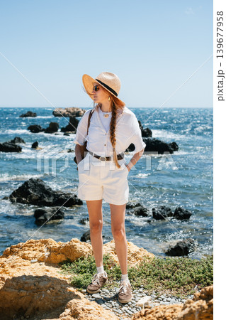 Woman in white outfit and straw hat enjoying scenic ocean view on rocky coastline Woman in white outfit and straw hat enjoying scenic ocean view on rocky coastline 139677958