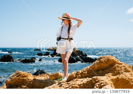 Woman exploring rocky seaside in summer outfit and hat 139677959