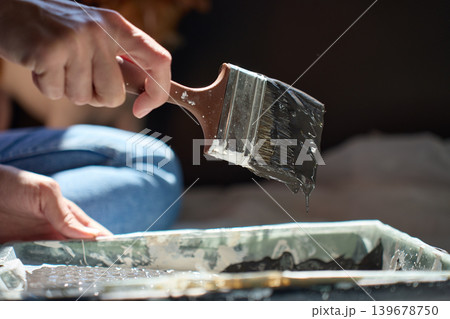 Person holds paintbrush with dark paint dripping over tray on protective sheet Person holds paintbrush with dark paint dripping over tray on protective sheet 139678750