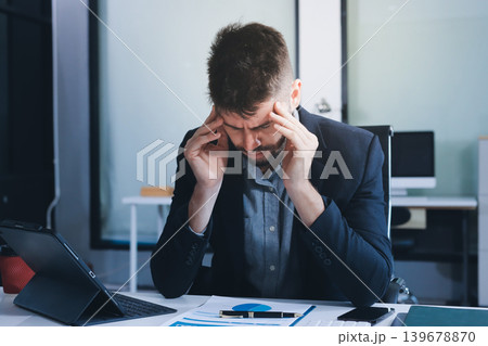 Tired worker. A tired businessman works in the evening. A man in office clothes is holding his temples. Problems in business. The businessman is trying to solve the problems that have arisen. 139678870