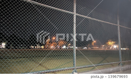 Nighttime view of an empty sports arena captured through a prominent chain link fence. Bright stadium lights illuminate the green grass field, surrounding city trees. Distant houses emit warm orange Nighttime view of an empty sports arena captured through a prominent chain link fence. Bright stadium lights illuminate the green grass field, surrounding city trees. Distant houses emit warm orange 139679693