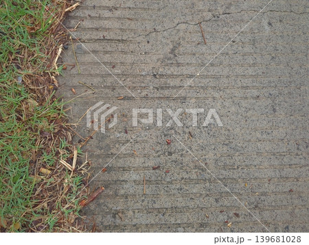 View of a concrete path with horizontal lines and a section of green grass along the left edge. The contrast between the rigid, man-made surface and the unruly, organic grass creates a natural divide. 139681028