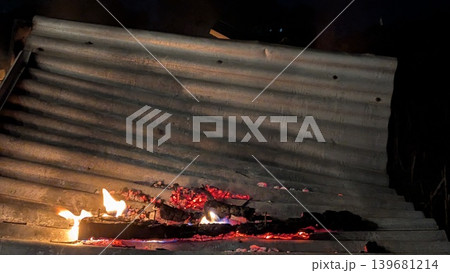 A fire blazes on a corrugated metal roof, casting a fiery glow and revealing glowing embers. The long exposure captures the motion of the flames and rising smoke against the dark night. A fire blazes on a corrugated metal roof, casting a fiery glow and revealing glowing embers. The long exposure captures the motion of the flames and rising smoke against the dark night. 139681214