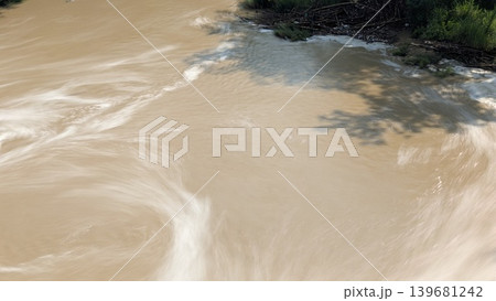 An artistic long exposure of a muddy brown river, showing silky white swirls of moving water, with a dark bank of greenery, foam, debris on the top edge, a shadow of an overhead object. 139681242