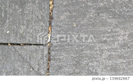A close-up, top-down view of a gray concrete surface made of interlocking, rectangular blocks. The pavement has visible cracks and a rough texture, with a thin line of dirt and leaves in the center. 139682667