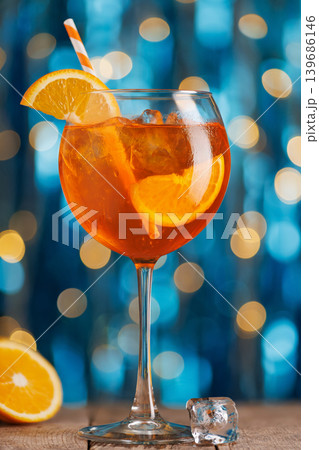 Aperol spritz cocktail with ice, orange slice garnish and striped straw on wooden table, bright blue bokeh background evoking festive summer refreshment at party Aperol spritz cocktail with ice, orange slice garnish and striped straw on wooden table, bright blue bokeh background evoking festive summer refreshment at party 139686146