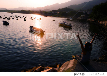 Woman enjoys sunset over tranquil bay with boats and distant mountains Woman enjoys sunset over tranquil bay with boats and distant mountains 139687364