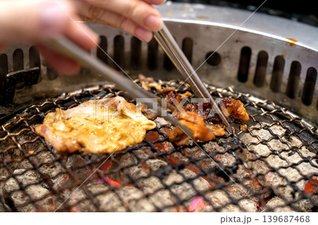 Close up of meat grilling on a charcoal barbecue grill with hands using metal chopsticks to turn the food. Traditional yakiniku or Korean BBQ dining experience with glowing embers and smoke. 139687468