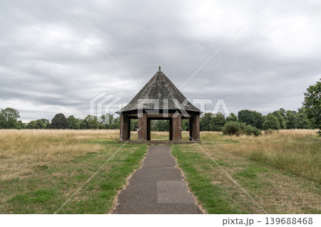 Open brick pavilion standing beside grassy fields under dramatic cloudy skies. Peaceful countryside scenery creates a calm outdoor landscape atmosphere. Open brick pavilion standing beside grassy fields under dramatic cloudy skies. Peaceful countryside scenery creates a calm outdoor landscape atmosphere. 139688468