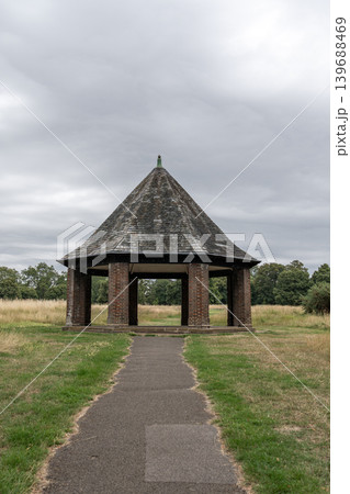 Open brick pavilion standing beside grassy fields under dramatic cloudy skies. Peaceful countryside scenery creates a calm outdoor landscape atmosphere. 139688469