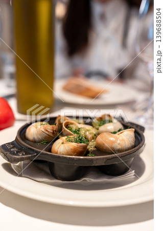 Cooked escargot in shells is topped with garlic herb butter on a black pan. White plates and drinks appear blurred in the restaurant background. 139688504
