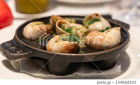 Overhead view shows escargot in shells with garlic herb butter served on black pans and white plates, surrounded by drinks and utensils in a restaurant dining setting. Overhead view shows escargot in shells with garlic herb butter served on black pans and white plates, surrounded by drinks and utensils in a restaurant dining setting. 139688505