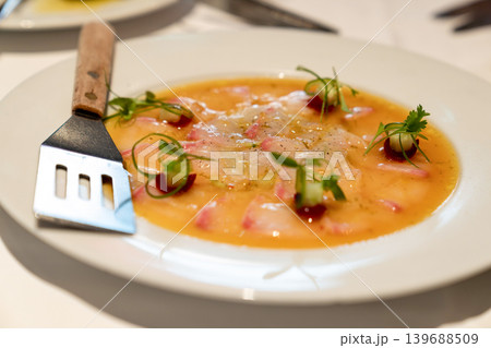 Thinly sliced fish served with citrus dressing, garnished with microgreens and decorative sauce dots. Plate and fork visible on white table. Thinly sliced fish served with citrus dressing, garnished with microgreens and decorative sauce dots. Plate and fork visible on white table. 139688509