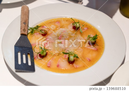 Thinly sliced fish served with citrus dressing, garnished with microgreens and decorative sauce dots. Plate and fork visible on white table. 139688510
