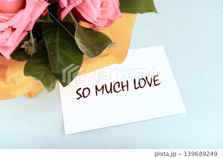 A vibrant bouquet of pink roses and green foliage wrapped in orange paper sits beside a white card bearing the heartfelt message SO MUCH LOVE on a gentle blue background A vibrant bouquet of pink roses and green foliage wrapped in orange paper sits beside a white card bearing the heartfelt message SO MUCH LOVE on a gentle blue background 139689249