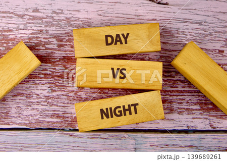 Three wooden blocks spell DAY VS NIGHT, stacked on a faded red and white wooden background. More blocks are scattered, adding to this conceptual representation of comparison 139689261