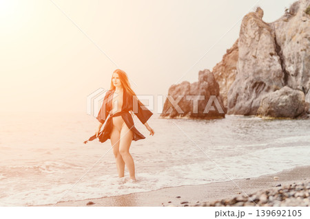 Woman beach swimsuit walks along the shoreline by the ocean near large rocks at sunset Woman beach swimsuit walks along the shoreline by the ocean near large rocks at sunset 139692105