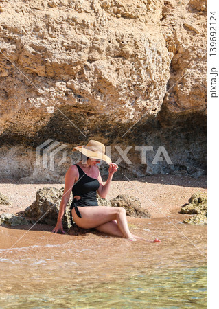 Young woman in a black swimsuit, hat, and sunglasses posing for the photographer against a stunning rocky seaside backdrop, capturing the essence of summer and beach vibes 139692124