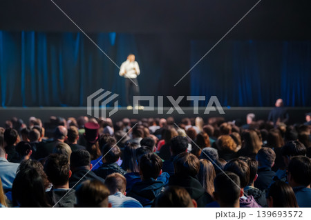 Large audience watching speaker on stage during conference presentation with dim blue curtains 139693572