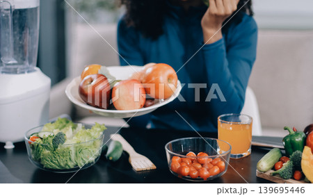 Young nutritionist showing bell peppers during an online video call consulting about healthy eating, sitting at home at a table with vegetables, laptop and measuring tape 139695446