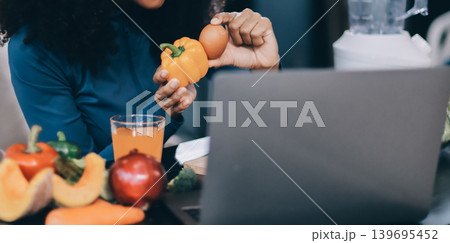 Young nutritionist showing bell peppers during an online video call consulting about healthy eating, sitting at home at a table with vegetables, laptop and measuring tape Young nutritionist showing bell peppers during an online video call consulting about healthy eating, sitting at home at a table with vegetables, laptop and measuring tape 139695452