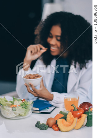 Young nutritionist showing bell peppers during an online video call consulting about healthy eating, sitting at home at a table with vegetables, laptop and measuring tape Young nutritionist showing bell peppers during an online video call consulting about healthy eating, sitting at home at a table with vegetables, laptop and measuring tape 139696959