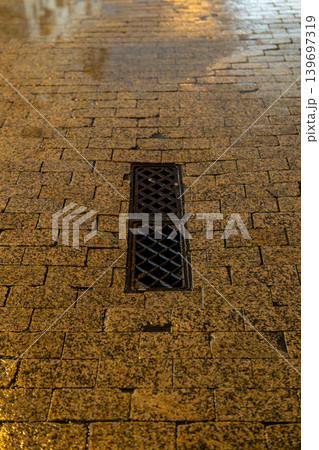 Wet Cobblestone Street With Metal Drain Grate at Night Reflective Urban Pavement Texture 139697319