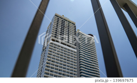 Low-angle view of a modern high-rise building framed by metal bars against a clear blue sky, highlighting urban architecture, perspective, and city living 139697675