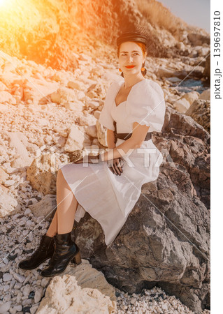 Woman white dress sunset elegant lady sitting on coastal rocks during golden hour for a summer lifestyle portrait Woman white dress sunset elegant lady sitting on coastal rocks during golden hour for a summer lifestyle portrait 139697810