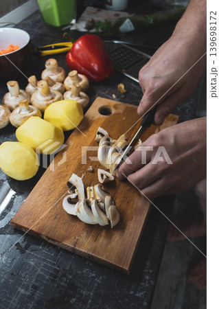 Dinner preparing process. Man's hands chopping vegetables on cutting board. man preparing vegetable soup. Peeled potatoes, carrot with mushrooms and red bell pepper on the table. Dinner preparing process. Man's hands chopping vegetables on cutting board. man preparing vegetable soup. Peeled potatoes, carrot with mushrooms and red bell pepper on the table. 139698172