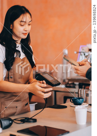 Asian senior female barista adult receiving mobile payment from customer using smartphone at cafe demonstrating modern cashless transaction in cozy local family coffee shop Asian senior female barista adult receiving mobile payment from customer using smartphone at cafe demonstrating modern cashless transaction in cozy local family coffee shop 139698824