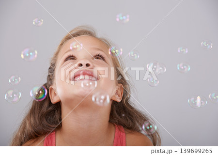 Smile, little girl and bubbles in studio for fun, excited and enjoy games with soap liquid magic. Child, happy and learning to play with bubble, natural and wellness in looking up by gray background Smile, little girl and bubbles in studio for fun, excited and enjoy games with soap liquid magic. Child, happy and learning to play with bubble, natural and wellness in looking up by gray background 139699265