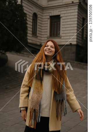 Smiling red-haired girl walks down a street in a city during daylight wearing a coat and scarf Smiling red-haired girl walks down a street in a city during daylight wearing a coat and scarf 139700506