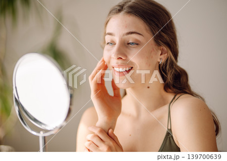 Young woman with problem skin looking into mirror. Concept of acne therapy, scars Young woman with problem skin looking into mirror. Concept of acne therapy, scars 139700639