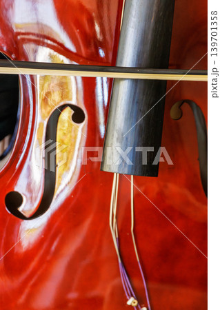 Close up of a glossy contrabass body with f hole, bow, tailpiece, and loose detached strings hanging over the varnished wood. Close up of a glossy contrabass body with f hole, bow, tailpiece, and loose detached strings hanging over the varnished wood. 139701358
