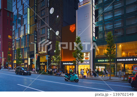 Tokyo, Japan - Oct 14 2024, panoramic view of main street of Ginza district, tall glass facades of skyscrapers along which people walk, cars drive along road, in evening with illumination, Tokyo Japan 139702149