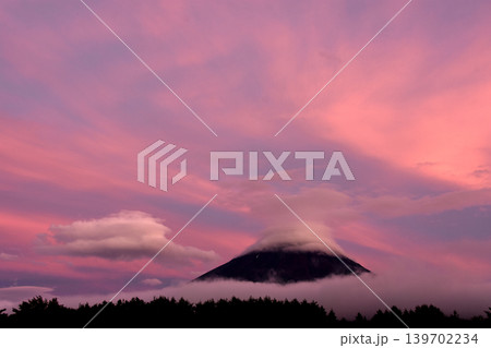 夏の夕焼けに染まる高層の薄雲をバックに白い笠雲をかぶった漆黒の富士山と横に浮かぶ二段の吊るし雲 139702234