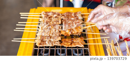 The shot of a cook grill Moo Ping or grilled pork skewers in Thai style on the yellow electric barbeque grill at the restaurant. The shot of a cook grill Moo Ping or grilled pork skewers in Thai style on the yellow electric barbeque grill at the restaurant. 139703729