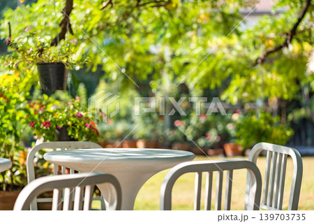 The outdoor furniture set, a white round table with seats, placed in the peaceful and shady garden. Shadow of branches and leaves project on outdoor furniture. 139703735