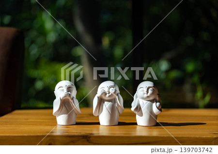 The white ceramic statue of three little monk See, Hear, Speak no evil set for home decoration placed on the wooden table over the black wall. The white ceramic statue of three little monk See, Hear, Speak no evil set for home decoration placed on the wooden table over the black wall. 139703742
