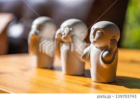 The white ceramic statue of three little monk See, Hear, Speak no evil set for home decoration placed on the wooden table over the black wall. The white ceramic statue of three little monk See, Hear, Speak no evil set for home decoration placed on the wooden table over the black wall. 139703743