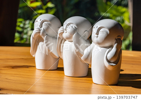 Three white monk figurines inspired by the Three Wise Monkeys philosophy are displayed as minimal zen decoration on a wooden tabletop with peaceful spiritual atmosphere. Three white monk figurines inspired by the Three Wise Monkeys philosophy are displayed as minimal zen decoration on a wooden tabletop with peaceful spiritual atmosphere. 139703747