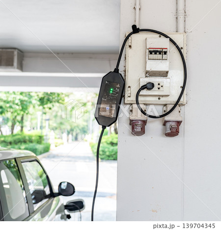 Portable electric vehicle charger connected to wall power outlet in parking area, representing sustainable transportation and clean energy technology. Portable electric vehicle charger connected to wall power outlet in parking area, representing sustainable transportation and clean energy technology. 139704345