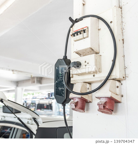 Portable electric vehicle charger connected to wall power outlet in parking area, representing sustainable transportation and clean energy technology. Portable electric vehicle charger connected to wall power outlet in parking area, representing sustainable transportation and clean energy technology. 139704347