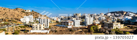 High angle panoramic view of the buildings and mountains in Qarn Dhabi, Al Bahah, Saudi Arabia, showing the urban development and arid hills under a clear blue sky. 139705027