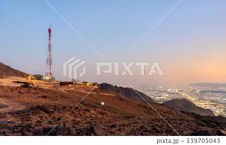A scenic panoramic view of Madinah city lights from the rocky peak of Jabal Uhud at dusk with a telecommunications tower in the foreground. A scenic panoramic view of Madinah city lights from the rocky peak of Jabal Uhud at dusk with a telecommunications tower in the foreground. 139705043