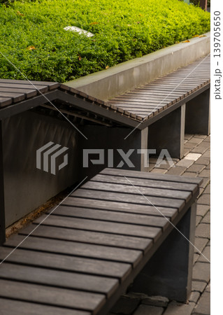 Modern outdoor wooden benches arranged beside landscaped greenery in an urban public space with clean architectural design. Modern outdoor wooden benches arranged beside landscaped greenery in an urban public space with clean architectural design. 139705650