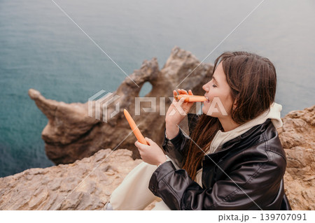 Woman eating carrots outdoors on a rocky cliff overlooking the sea while wearing a leather jacket during a hike Woman eating carrots outdoors on a rocky cliff overlooking the sea while wearing a leather jacket during a hike 139707091